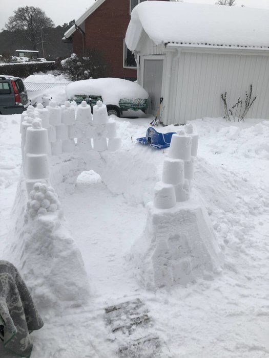 Hemmagjord snöborg med torn och pelare i en snöig trädgård, med hus och snötäckt bil i bakgrunden.