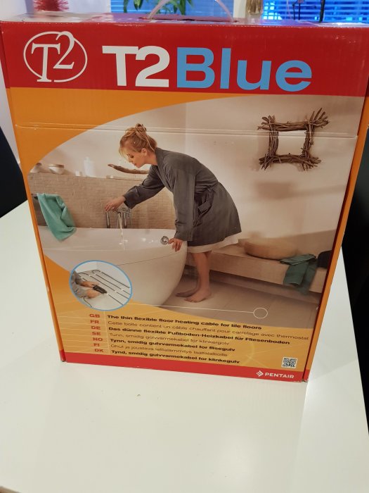 Förpackning av T2Blue golvvärme med bild av kvinna som justerar termostat vid handfatet.
