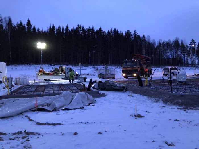 Arbetare förbereder gjutning vid gryning med betongbil och betongpump på en byggarbetsplats med snötäckt mark.