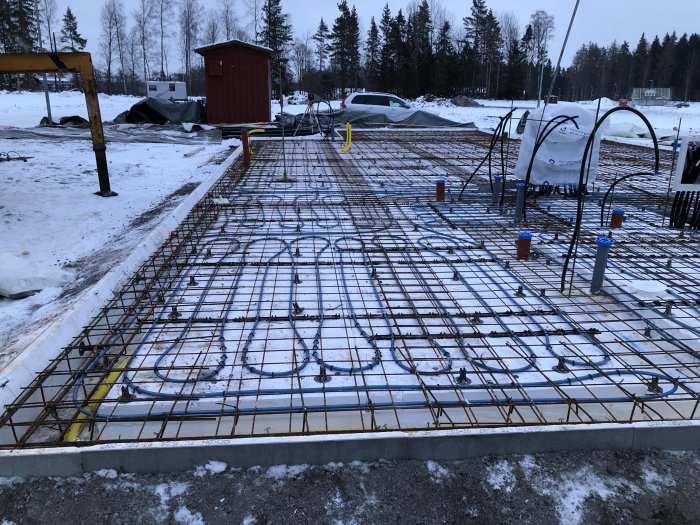 Armeringsjärn och rörinstallationer på en byggplats med snötäckt mark och presenningar i bakgrunden.