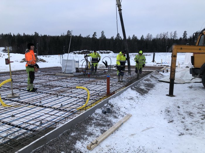 Arbetare förbereder för gjutning av betongplatta på vinterbygge med armeringsjärn och betongpump i förgrunden.