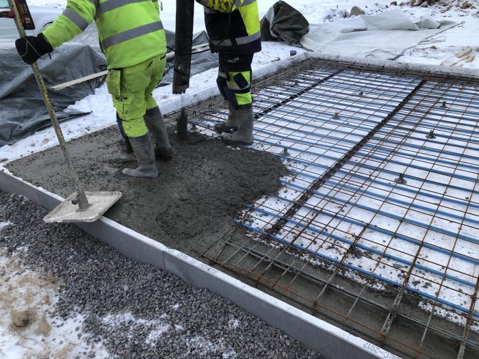 Två arbetare i säkerhetskläder gjuter betong för en byggplatta utomhus med synlig armering och snötäckt mark.