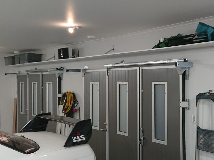 Garage med uppfällda hyllor, skjutdörrar och del av en bil med WRC-märke.