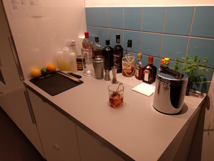 Kök med ny bänkskiva och blå kakel, diverse drycker och citrusfrukter, cocktailshaker och ishink.