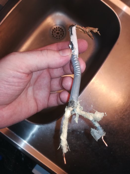 Hand håller gammal elledning med synliga koppartrådar och slitet isoleringsmaterial som påminner om papper eller tyg.
