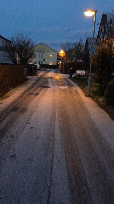 Tunn snötäckning på en gata i bostadsområde med gatlyktor tända i skymningen.