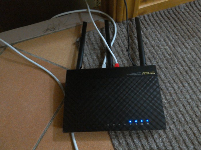 En trådlös ASUS-router med fyra antenn och upplysta statuslampor på en matta, nära ett skrivbord.