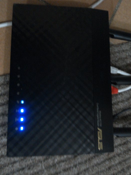 Router från ASUS med tända ljusindikatorer på en matta och kablar i bakgrunden.