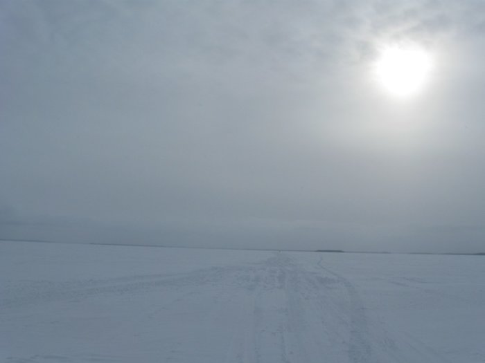 Övergiven bilväg överfrusen med snö och is under disig himmel med svag solsken.