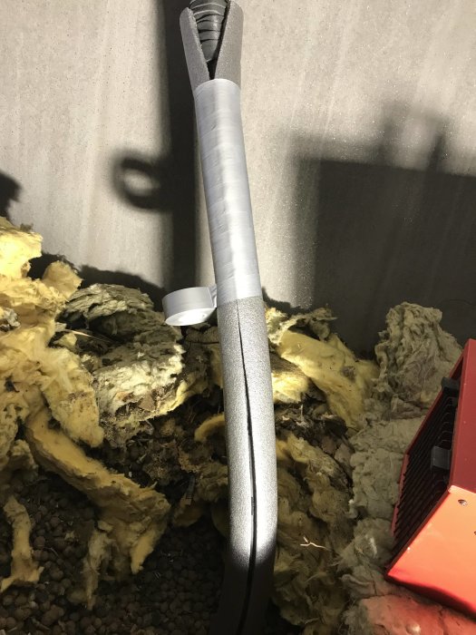 Ett isolerat PEM-rör med tejp och polyetenisolering omgiven av gammal mineralull och grus på marken.