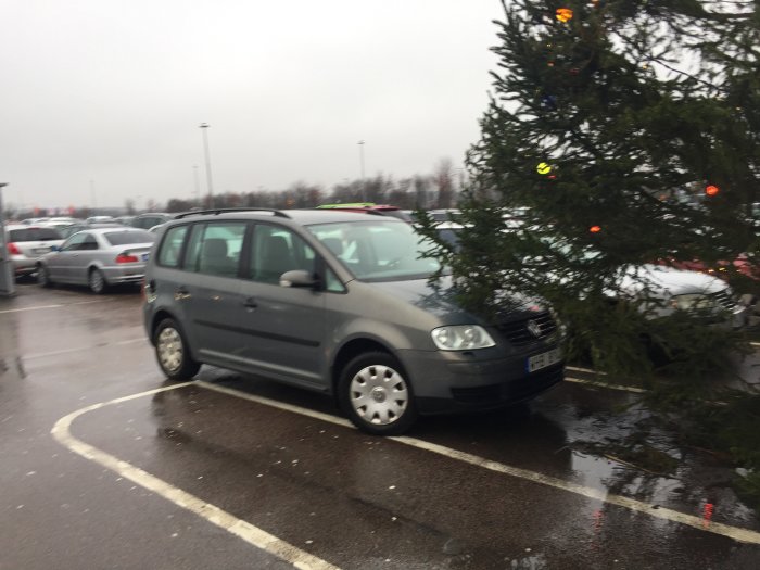 Grå bil parkerad bredvid en stor julgran mitt på en parkeringsplats.