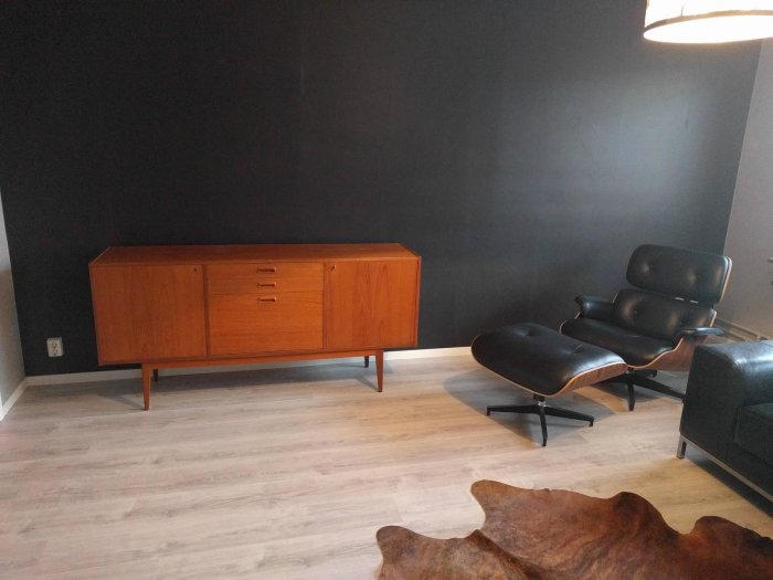 Vardagsrum med mörk fondvägg, mid century-stil sideboard och fåtölj, samt synligt original ekparkettgolv.