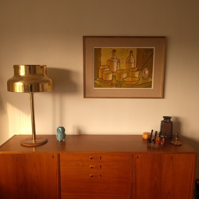 Mid century-stil vardagsrum med träbyrå, gyllene bordlampa och konstverk av föremål i brun ram på en vägg.