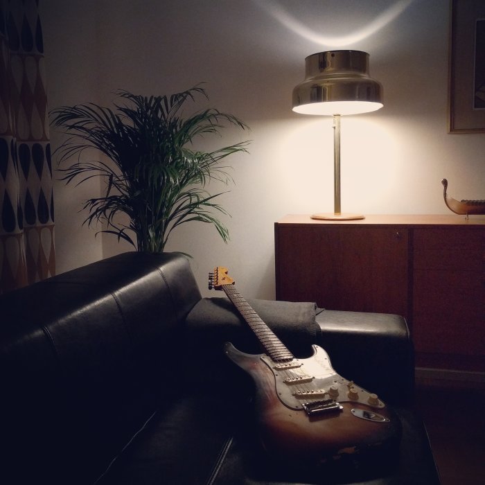 Inredd hörna av vardagsrum i mid century-stil med svart soffa, golvstående lampa, träbyrå och elgitarr.