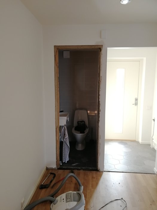Ett badrum sett genom en dörröppning utan karm med verktyg och en dammsugare på golvet i förgrunden.