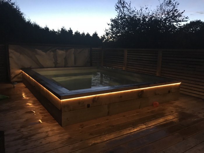 Pool med ljustemaslinga på kvällen, trätrall, staket och växter i bakgrunden.