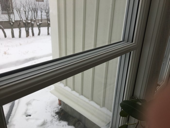 Närbild på ett fönster med aluminium spröjs utomhus, snötäckt mark syns i bakgrunden.