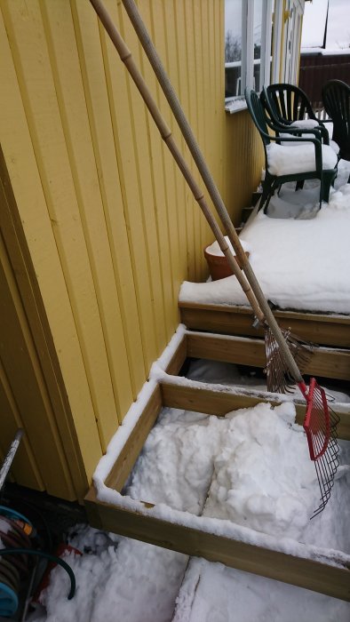 Snöbelagd trätrappa med en räfsa lutad mot ett gult trähus.