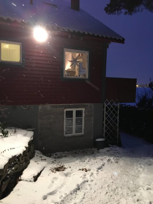 Rödmålat hus med nytt fönster utan spröjs i snötäckt omgivning vid skymning.