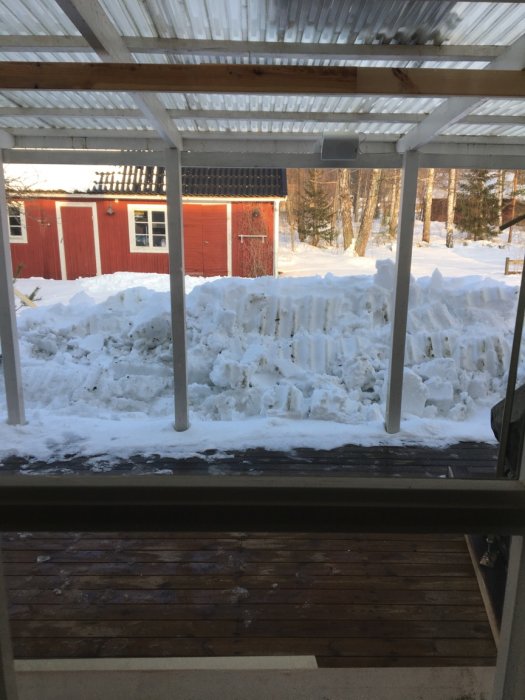 Vy från en altan med tak som visar en stor hög av skottad snö framför ett rött hus med vita knutar i vintersol.