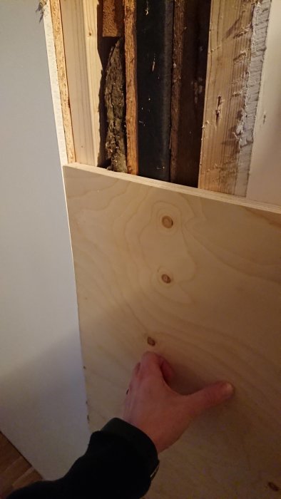 Hand håller ett stycke plywood vid en gammal väggöppning under renovering.