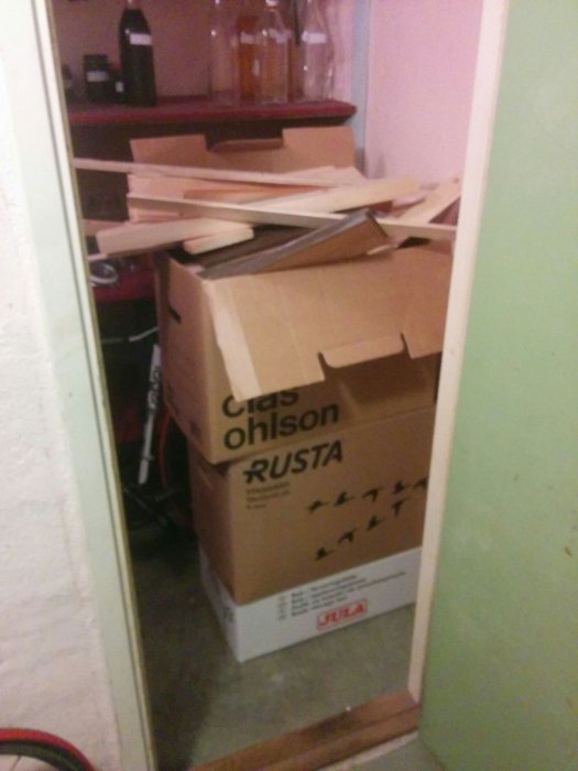 Rörig arbetsplats med en kartong märkt 'Clas Ohlson' och 'Rusta', överfull med virkesbitar.