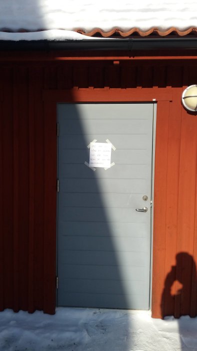 Stängd grå ytterdörr på en röd träpanel med snö på marken, solskugga och ett vitt pappersark i mitten av dörren.