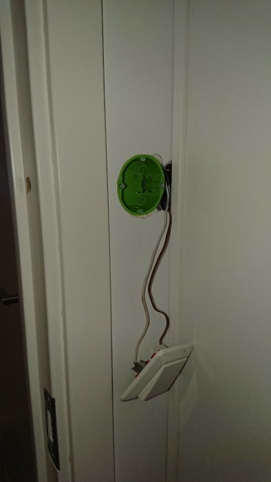 Grön installationsdosa i vägg med utstickande elkablar, bredvid en öppen strömbrytare.