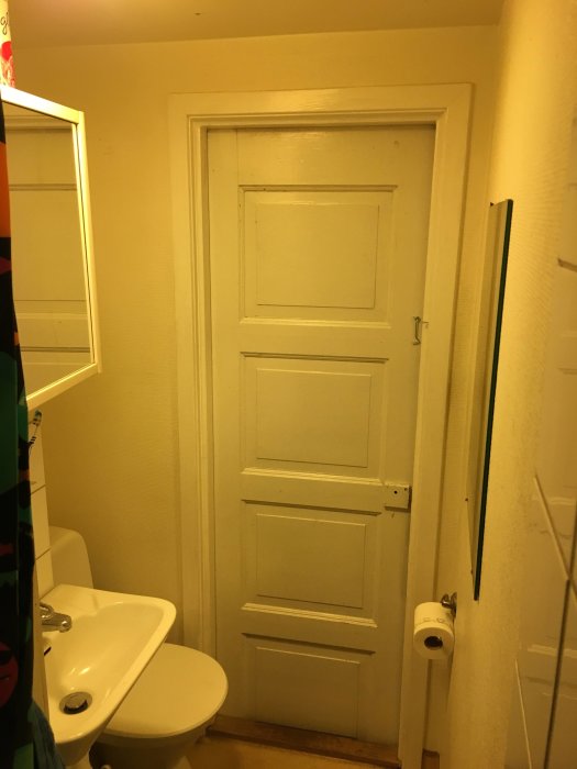 Ommålat badrum med beige väggar och vit dörr, före renovering, inkluderar handfat och toalett.