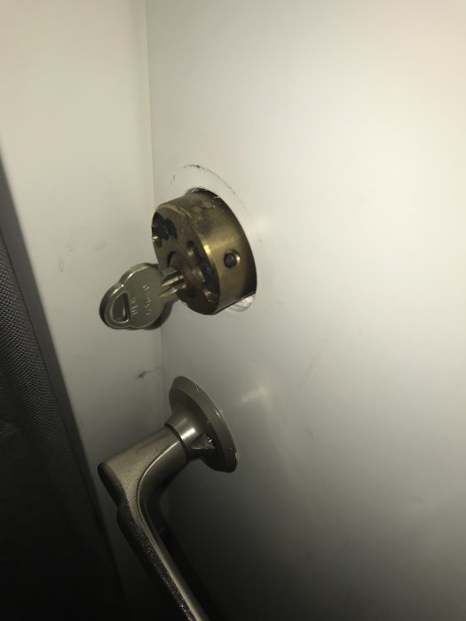 Nyckel i mässingscylinder med fastsittande ring på dörr, bredvid dörrhandtag.