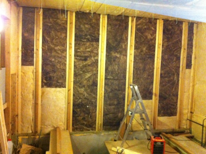 Vägg under renovering med synlig isolering och uppsatta vertikala läkter redo för montage av dubbla lager gips för ljudisolering.