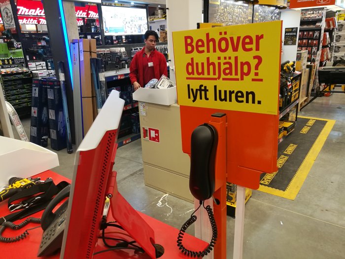 Kund i byggvaruhus väntar vid en kundtjänststation med skylten "Behöver du hjälp? lyft luren".