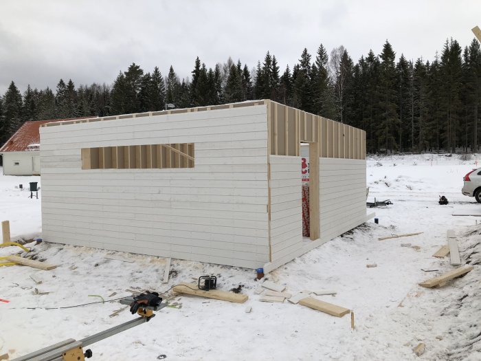 Halvfärdigt garage med paneling, spikläkt och utomhusgips mot en skogsbakgrund i vinterscen.