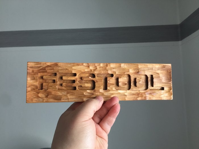 En hand håller en träplakett med utskuren text "FESTOOL" mot en grå bakgrund.