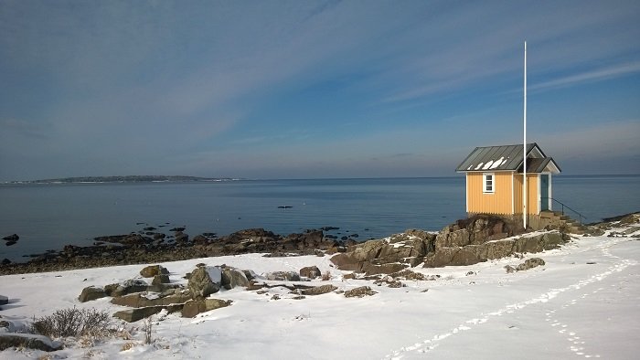 Snötäckt kustlandskap i Skåne med litet hus vid havet under en klarblå himmel.