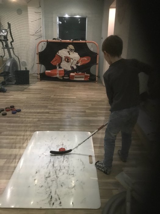 Barn spelar hockey inomhus med klubba och boll mot en målvaktstavla.