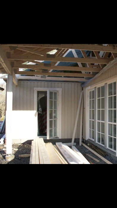 Underkonstruktion av en altan med taksparrar och en partiell vägg, intill ett hus med fönster och en öppen dörr.