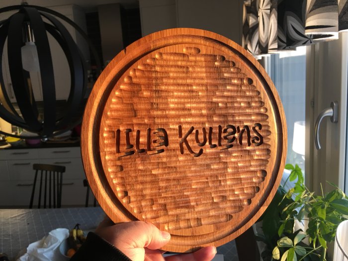 En hand håller en träplatta med fräst text "Illa Kulens" och mönstrad kant.