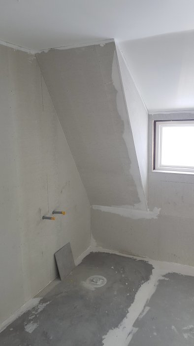 Obehandlat badrum med snedtak, gipsvägg och rörutstick vid fönster.