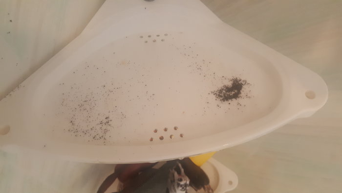 Vit plastdel med svarta partiklar och grus, möjliga manganutfällningar, spridda ojämnt över ytan.