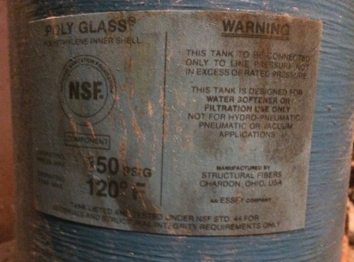 Närbild av sliten märklapp på en blå glasfiberfiltertank med teknisk information och varningar.