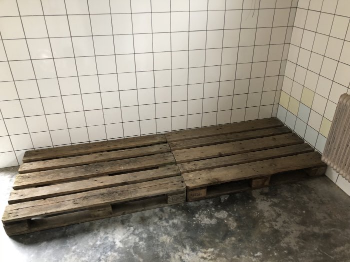 Träpallar staplade i ett hörn av en källare med vita kakelväggar och betonggolv, förberedelse för golvläggning.