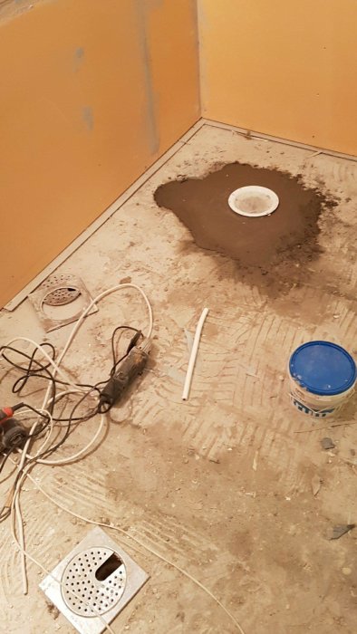 Golv under renovering med en golvbrunn och kakelfärdigt vägg samt verktyg och byggmaterial.