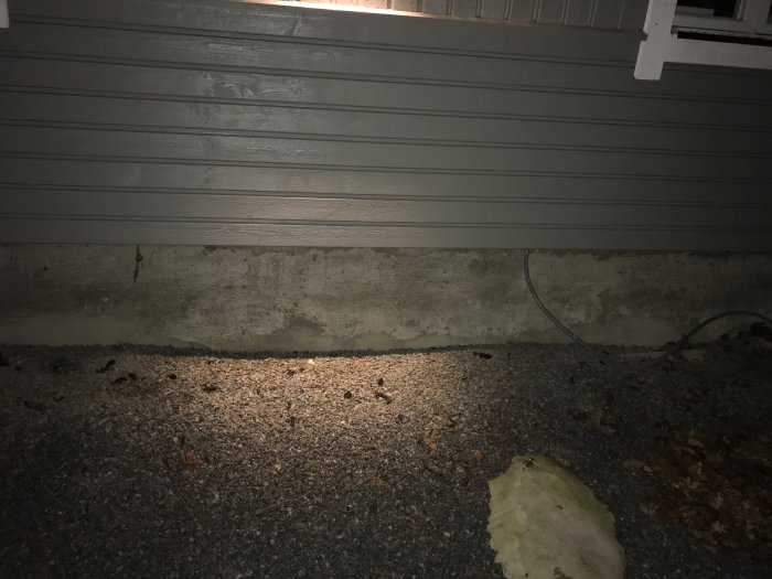 Husvägg med träpanel ovanför en grå betongplatta, med synliga kablar och grusig mark nedanför.
