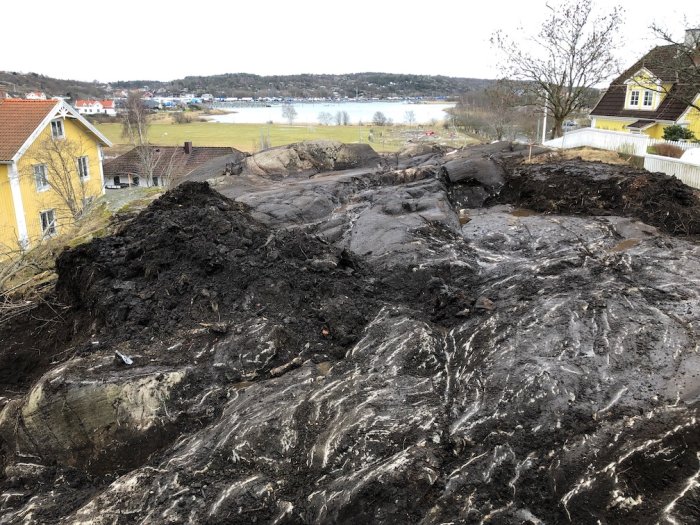 Markarbete på en lutande tomt med grävd jord och berg, med vatten och hus i bakgrunden.