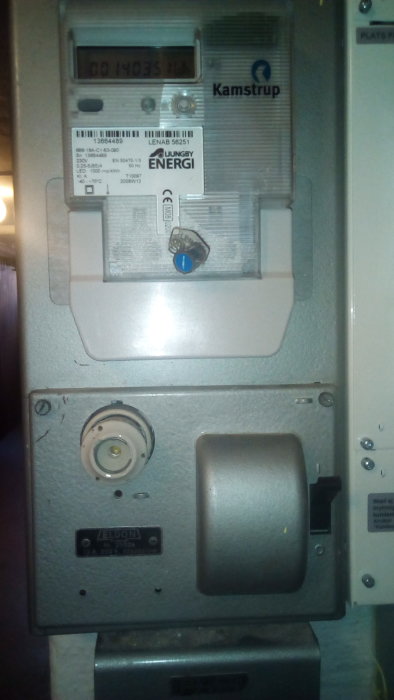 Eldgammal elektrisk mätare med svarta skyltar och otydligt serienummer på ett blågrått skåp.
