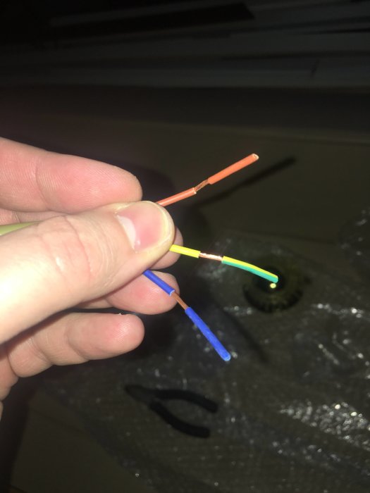 En hand som håller en brun, blå och grön/gul elkabel framför ett öppet hål i taket.