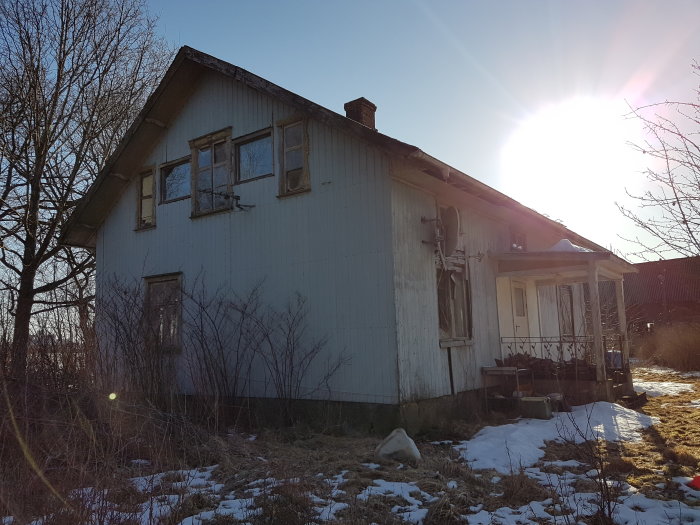 Tvåvånings trästuga i behov av renovering med solnedgång i bakgrunden, något snö på marken.