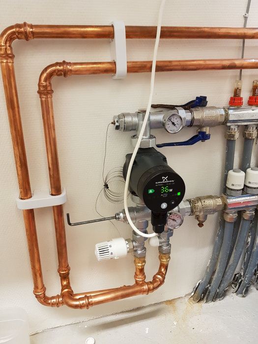 Kopparledningar och cirkulationspump i ett värmesystem med justerbar flödesventil och manometer.
