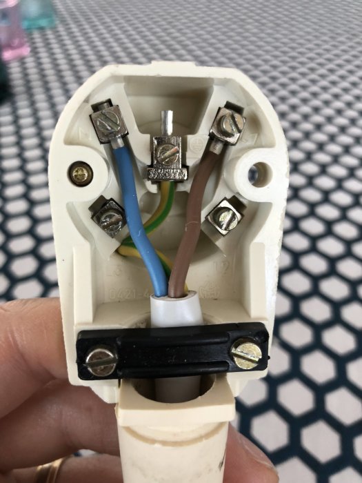 Hand som håller en öppnad stickkontakt med synliga anslutningar och färgkodade kablar mot ett mönstrat underlag.